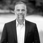 A man with short gray hair and a beard wearing a dark suit jacket and light checkered shirt, standing outdoors with a blurred background.