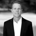 A man in a business suit poses for a formal portrait against a blurred outdoor background.