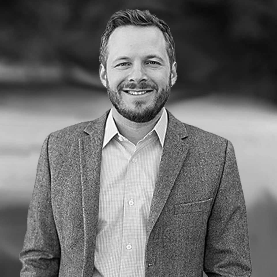 A man with a short beard and mustache is wearing a checked shirt and a blazer, smiling while standing outdoors in front of a blurred background.